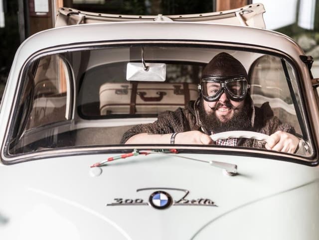 Een man zit lachend achter het stuur van een oude auto. Hij draagt een bijzondere bril en een helm, wat retro oogt.