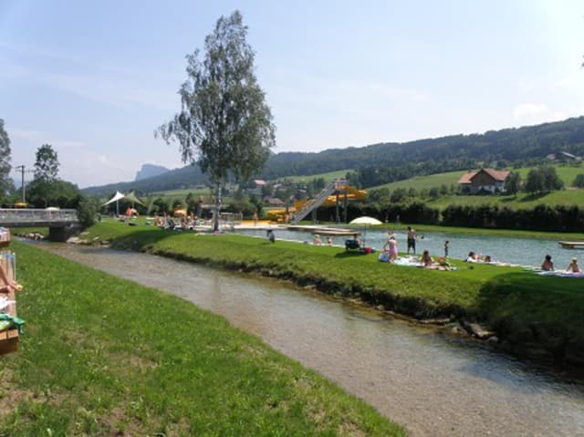 Een rustige rivier met groene weiden en bomen. Op de achtergrond genieten mensen van het water en de zon.