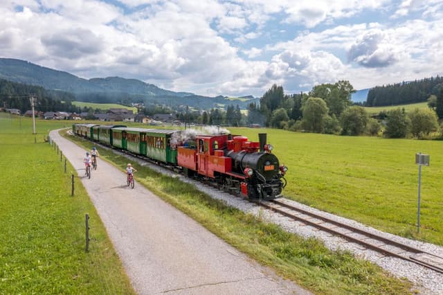 Una locomotiva a vapore attraversa un paesaggio verde. Ciclisti percorrono il sentiero accanto ai binari.