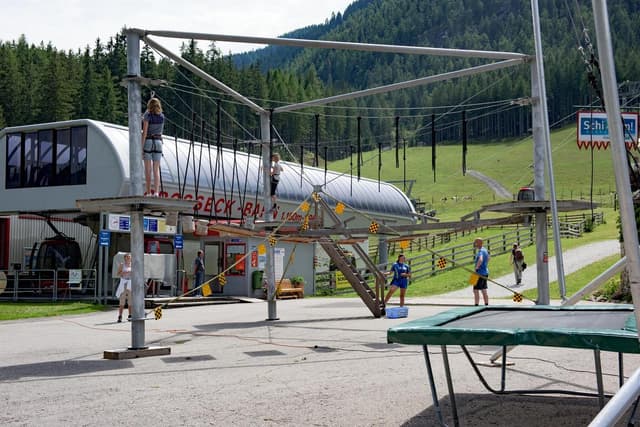 Park linowy z mostami linowymi i elementami wspinaczkowymi w alpejskim otoczeniu. W tle widać drzewa i budynek.