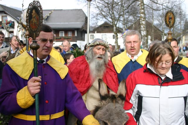 Un gruppo di persone in abiti da festa partecipa a una processione. In primo piano si trova un uomo anziano con la barba e un asino.