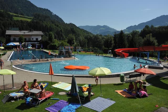 A swimming pool with a large expanse of water and a slide, surrounded by green meadows. Sun loungers and parasols invite relaxation.