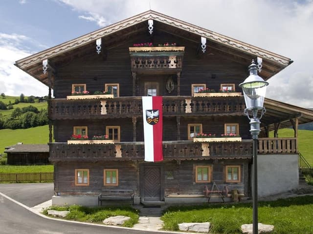 Una casa tradizionale in legno in stile alpino con balconi e un grande stemma. Davanti alla casa si trova un lampione, circondato da un paesaggio verde.