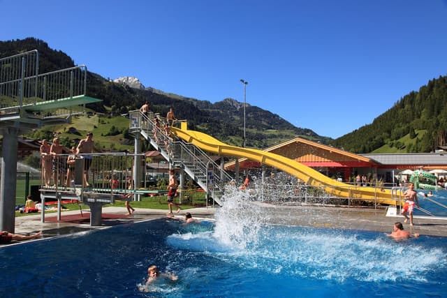 Basen ze zjeżdżalnią wodną i trampoliną do skoków. Wiele osób korzysta z dnia w wodzie i na słońcu.