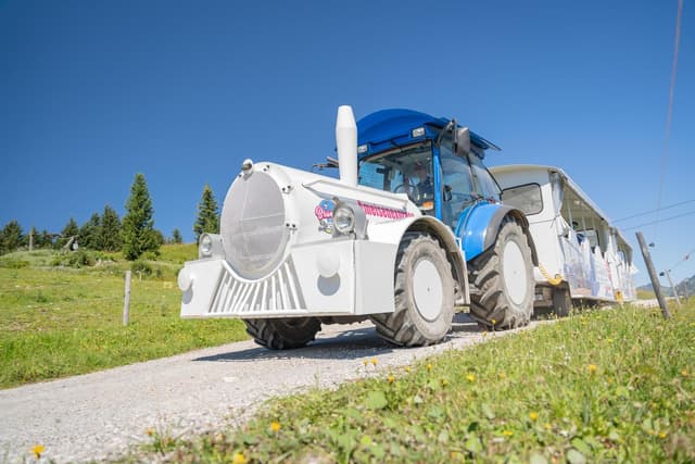 Neobvyklý traktor táhne vůz po štěrkové cestě. V pozadí jsou vidět zelené louky a stromy.
