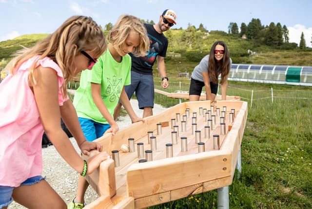 En gruppe børn og voksne leger udendørs ved et stort legeredskab af træ. Omgivelserne er grønne og bakkede, ideelle til aktiviteter i naturen.