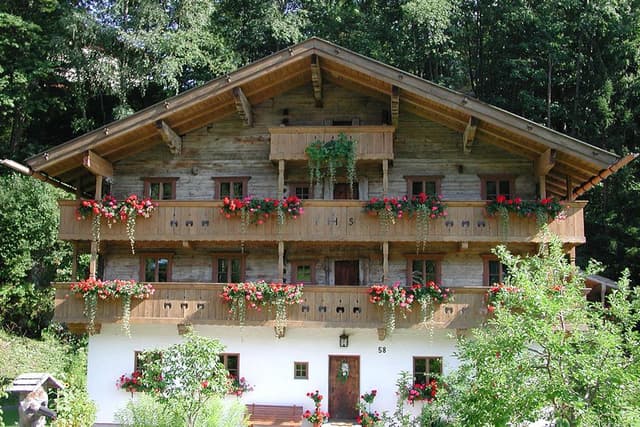 Tradiční dřevěný dům s kvetoucími truhlíky na balkonech. Obklopený zelenými stromy a jasnou oblohou.