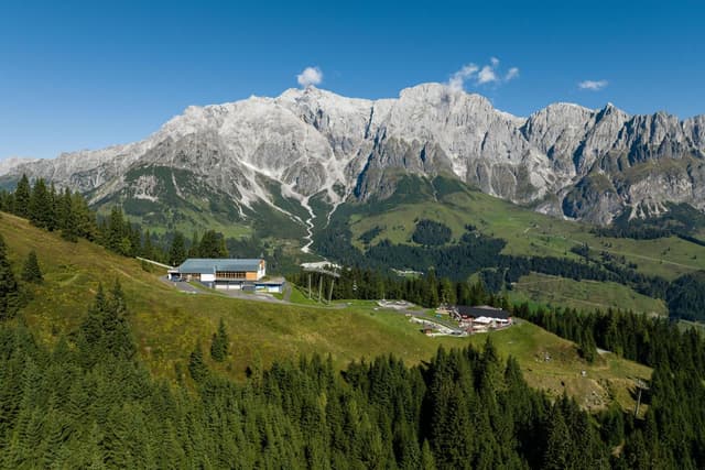 Malebná horská krajina se zasněženými vrcholy a zelenými loukami. V popředí stojí moderní budova obklopená stromy.