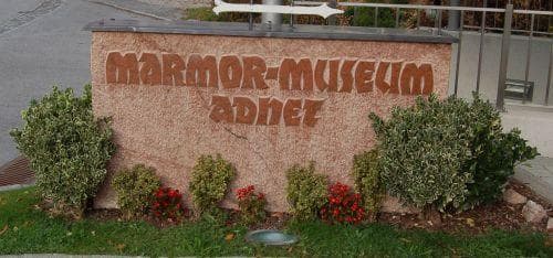 Kamenná cedule s nápisem „Marmor-Museum Adnet“. Zelené keře a červené květiny zdobí okolí.