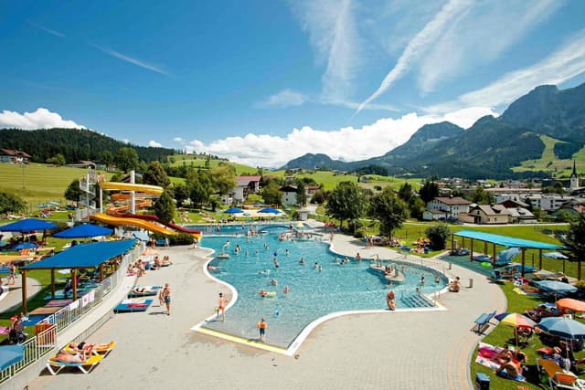 A swimming pool in a picturesque landscape with mountains in the background. Many visitors enjoy the water on a sunny day.