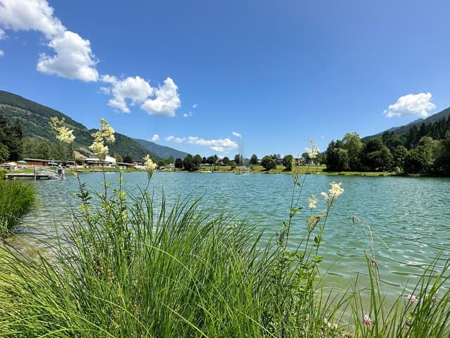 Klidné jezero obklopené zelenými loukami a horami. Obloha je jasná a slunečná s několika bílými mraky.