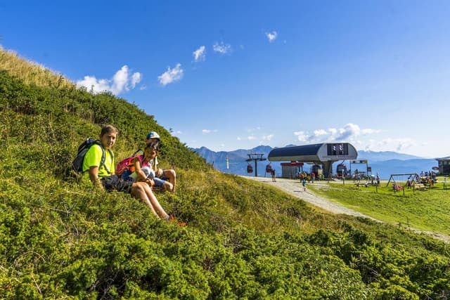 Twee kinderen zitten op een groene weide en genieten van een zonnige dag in de bergen. Op de achtergrond zijn een kabelbaan en het majestueuze landschap zichtbaar.