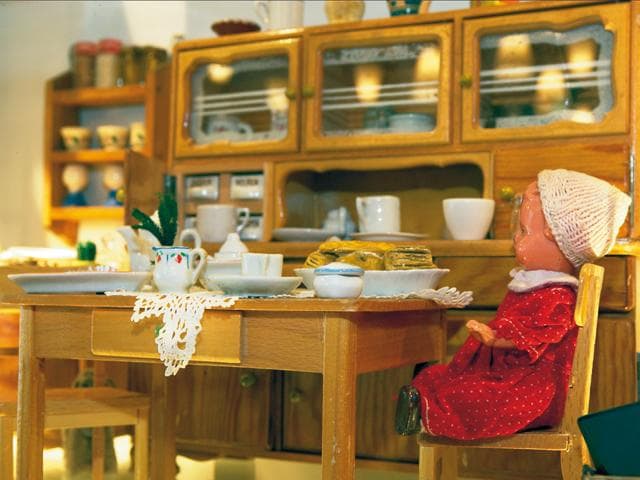 A cosy table in a doll's kitchen with laid-out crockery and a doll in a red dress. In the background shelves with crockery and foodstuffs are visible.