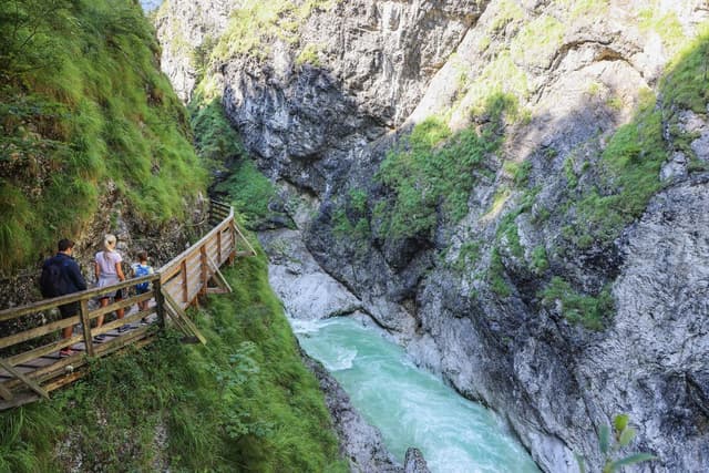 A picturesque gorge with green grass and steep rock faces. A narrow wooden walkway runs alongside the clear, flowing water.