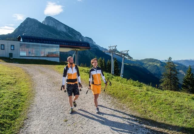 Dva turisté jdou po cestě v horách, v pozadí lyžařský vlek. Krajina je zelená, s vysokými horami a jasnou oblohou.
