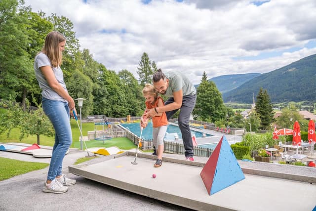 Rodina hraje minigolf v pěkném venkovním areálu. V pozadí jsou vidět zelené plochy a hory.