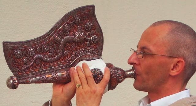 Muž fouká do bohatě zdobeného dechového nástroje. Detailní řemeslné zpracování nástroje je dobře vidět.