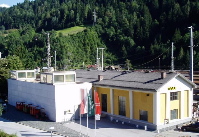 A modern building with a yellow façade and several aerials stands in a green mountain landscape. There are also some flags and orange containers in the foreground.