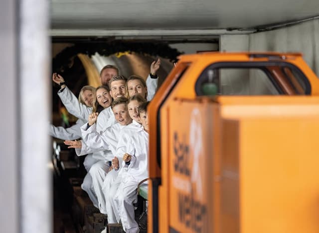 Skupina dětí v bílých overalech sedí na lavici a nadšeně ukazuje směrem k fotoaparátu. V popředí je vidět oranžový vozík.