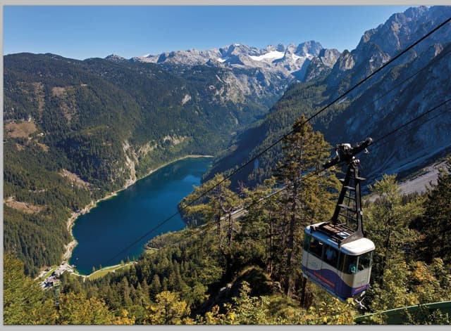 A cable car ride with a view of a clear lake and majestic mountains. The landscape is green and lush, with snow-covered peaks in the background.