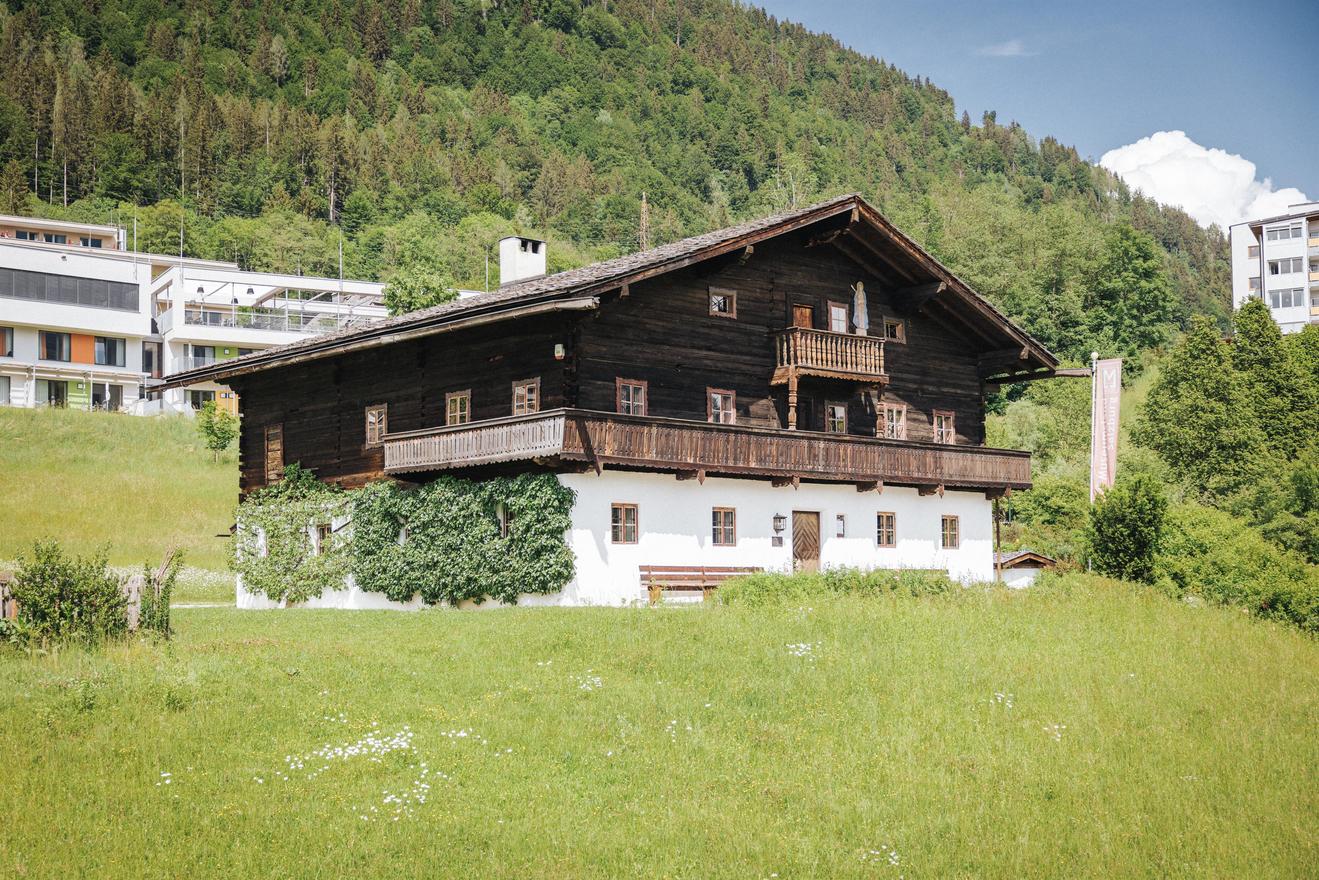 Tradiční dřevěný dům s balkonem stojí na zelené louce. V pozadí jsou vidět moderní budovy a zalesněný hřeben kopců.