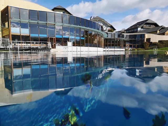 A modern building is reflected in the clear water of a pool. Surrounded by a calm landscape under a blue sky.