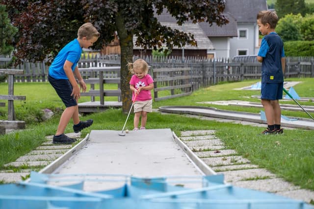 Drie kinderen spelen minigolf in een groen park. Een jongen staat klaar, terwijl een meisje met een golfstick mikt en de andere jongen toekijkt.