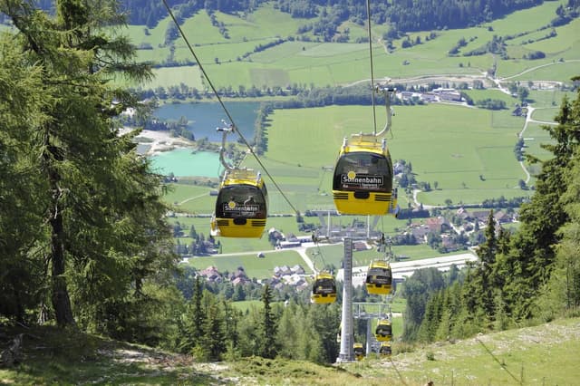 A cable car travels through a beautiful green forest. In the background, meadows and a lake are visible.