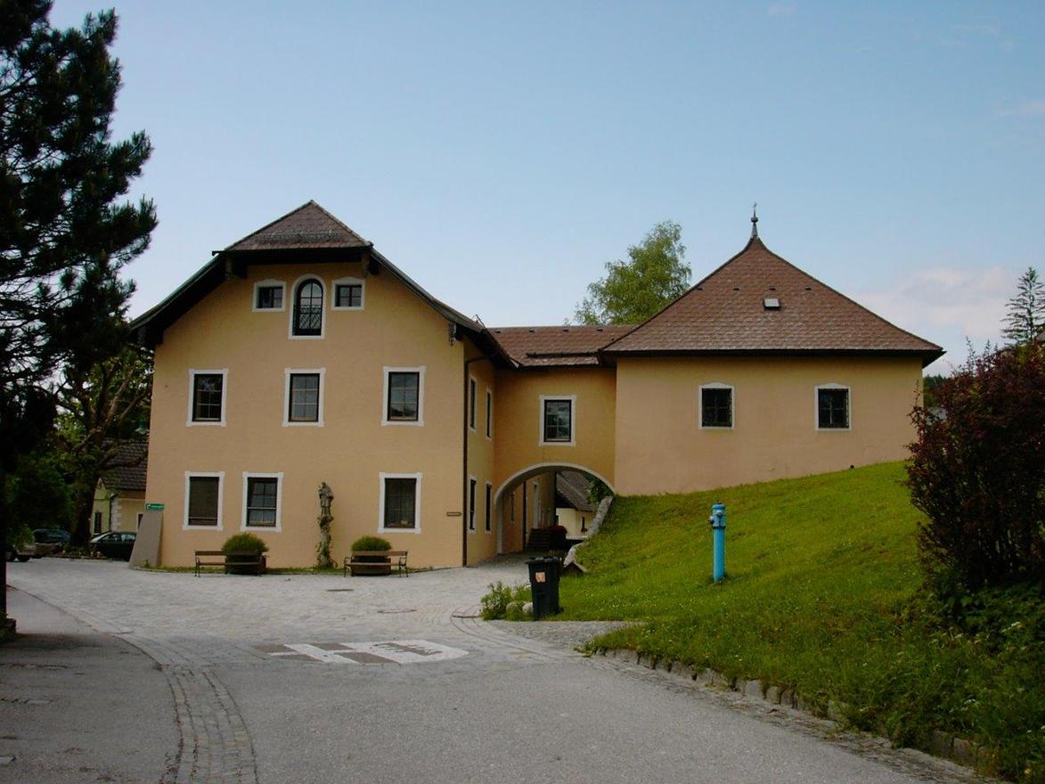 Žlutá budova s pěknou střechou a velkými okny. Stojí v klidném prostředí s množstvím zelené trávy a stromů.