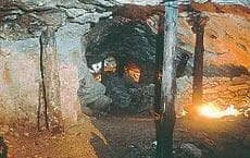 Podzemní štola s hrubými stěnami a dřevěnými trámy. V pozadí bliká slabé světlo.